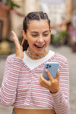 Diş teli takan beyaz bir kadın cep telefonu kullanıyor. Kutlama için cep telefonu kullanıyor. Açık havada piyango galibiyet ikramiyesi kazanıyor. Şehir caddesinde yürüyen mutlu kız. Yaşam Tarzları