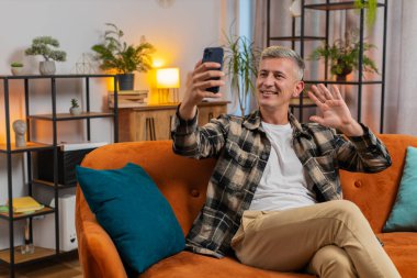 Beyaz saçlı mutlu beyaz adam akıllı telefondan selfie çekiyor, abonelerle internetten video görüşmesi yapıyor, sosyal medya için hikayeler kaydediyor. Genç adam kanepede oturuyor.