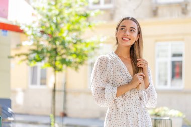 Düşünceli gülümseyen, düşünceli, mutlu, genç, yetişkin bir kadın. Çözüm üzerinde kafa yoran, soru soran, ilham veren açık havada rüya gören. Güneşli şehir sokağındaki güzel kız. Kasaba yaşam tarzları.