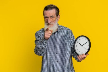 Senin sıran, acele et. Kıdemli yaşlı adam duvar ofis saati gösteriyor, tamam, başparmağınla kamerayı, reklamı, son teslim tarihini göster. Yetişkin büyükbaba sarı arka planda izole edilmiş.