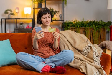 İnternetten alışveriş yapan mutlu genç bir kadın. Gülümseyen beyaz kız elinde bankacılık kredi kartı tutuyor evdeki koltukta mobil uygulama doğrulama sürecinde yer alıyor.