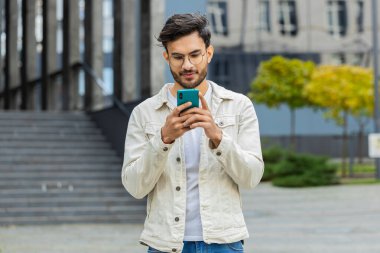 Genç gülümseyen Hintli adam akıllı telefon kullanıyor kısa mesajlar yazıyor, e-posta okuyor, dışarıda haberleri inceliyor. Arap Hindu adam şehir merkezindeki caddede duran mobil sosyal medya uygulamasını kullanıyor. İş adamları