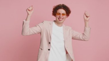 Beyaz, kıvırcık saçlı, müzik dinleyen, disko dansı yapan, kendini ifade eden eğlenceli elleri olan genç bir adam. Müzik uygulaması reklamı. Mutlu adam stüdyo pembe arka planda izole