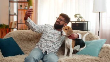 Hintli bir adam evde mutlu bir şekilde tazı köpeğiyle selfie çekiyor. Akıllı telefon kullanıyor ve birlikte oyun oynamaktan hoşlanıyor. Arap adam evcil hayvanıyla kanepede gülümser, neşe, sevgi, eğlence ve neşeli duygular gösterir.