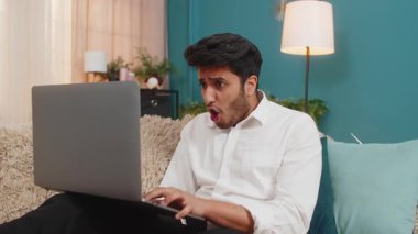 Hintli adam evde hayal kırıklığına uğramış hissediyor, online oyun kaybına tepki veriyor, hayal kırıklığı ve gerilim gösteriyor. Arap adam kanepede oturuyor, başarısız oyun denemesinden sonra stresli, dizüstü bilgisayara odaklanmış.