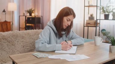 Not defterinde alışveriş listesi yapan beyaz bir kadın kanepede oturup makbuzlara bakıyor ve evdeki oturma odasında para sayıyor. Finansal maliyet veya bütçe için notlar hazırlayan kız
