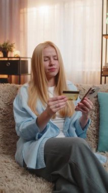 Evde genç bir kadın, akıllı telefon ve kredi kartıyla güvenli bir ödeme yapıyor. Kanepedeki kız, gün batımında sıcak oturma odasında son başarısına gülümseyen, onaylandıktan sonra rahatlar.