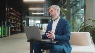 Modern ofis lobisinde dizüstü bilgisayarda çalışan orta yaşlı bir işadamı çalışanlarıyla online iletişim videosu görüşmesi yapıyor, patron. Bilgisayarın webcam görüntülerine bakan yönetici destek servisleri.