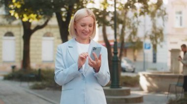 Kafkasyalı yetişkin iş kadını dışarıda cep telefonuyla internette sohbet ediyor. Mutlu gülümseyen orta yaşlı başarılı kadın cep telefonu kullanıyor. Şehir caddesinde SMS ile iletişim kuruyor.