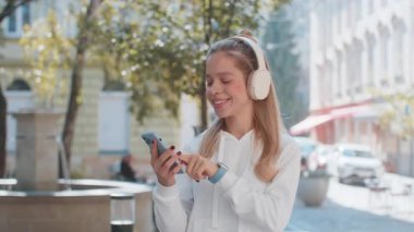 Gülümseyen, aşırı neşeli, kablosuz kulaklıklı kadın turist dışarıda dans eden akıllı telefondan müzik dinliyor. Şehir caddesinde günlük giysiler içinde yürüyen genç kız. Kasaba yaşam tarzları.