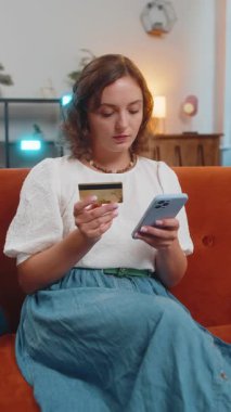 Para transfer ederken kredi kartı ve akıllı telefon kullanan mutlu genç bir kadın. Evin içinde alışveriş yaparken nakit para almadan yemek siparişi veriyor. Kanepede oturan kız. Dikey