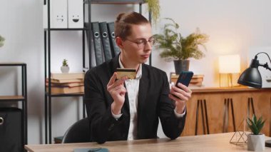 Kafkasyalı genç işadamı banka kartı ve akıllı telefonu kullanarak ev ofisinden online alışveriş yapıyor. İş anlaşması. Resmi takım elbiseli, cep telefonuyla gülümseyen serbest çalışan..