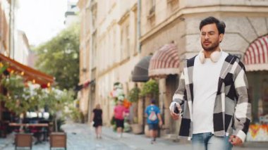Mutlu genç Hintli adam kablosuz kulaklık takıyor, açık havada akıllı telefondan en sevilen enerjik müziği dinliyor. Gülümseyen İspanyol turist şehir sokağının yanından geçiyor. Yaşam Tarzları.