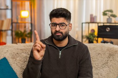 Evdeki Hintli adam, koltukta otururken ciddi bir yüz ifadesiyle HAYIR hareketi yapıyor, açıkça onaylamıyor. Arap adam, yoğun duygusal tepkiyle çatık kaşlı bir şekilde durumla olan anlaşmazlığını dile getiriyor.