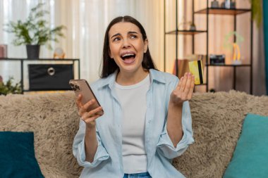 Evde genç bir kadın, engellenmiş hesap ya da hatalardan endişelenen akıllı telefondan kredi kartı bakiyesini endişeyle kontrol ediyor. Kanepedeki beyaz kız maddi sorunlar, kayıp ya da başarısız ödemeler konusunda vurguladı