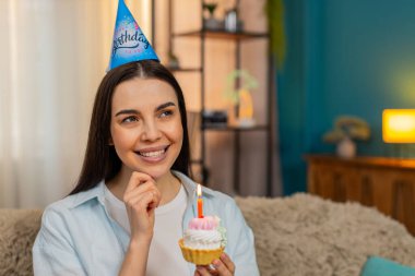 Evde elinde mumlarla mutlu bir şekilde gülümseyerek doğum günü pastası tutan genç bir kadın. Kanepedeki beyaz kız heyecanlı, duygusal olarak olumlu, neşeli bir ruh hali içinde, neşeli bir doğum günü kutlaması.