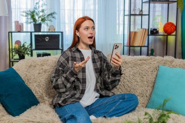 Evdeki genç kadın mobil uygulama kullanarak yeni bir dil öğreniyor ve kelimeleri odaklanma ve neşeyle yüksek sesle tekrarlıyor. Kanepedeki beyaz saçlı kız eğitim sürecinin tadını çıkarıyor. Telaffuz ve konuşma pratiği yapıyor.