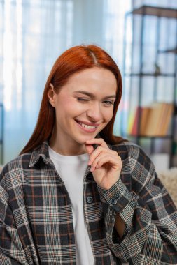 Genç bir kadın kameraya göz kırpıyor. Cilveli bir gülümsemeyle kendine güveni ve cazibesini gösteriyor. Kafkasyalı kızıl saçlı kız kanepenin üzerinde eğleniyor, duygularını ve kişisel çekiciliğini ifade ediyor.