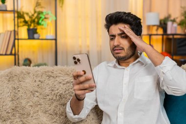 Evde oturan Hintli adam üzgün bir şekilde alnından akıllı telefon eline bakarak kötü haberler okuyor. Stresli hissediyor. Adam olumsuz mesaja tepki veriyor, endişeli, şok olmuş, beklenmedik bir bilgiyi işliyor.