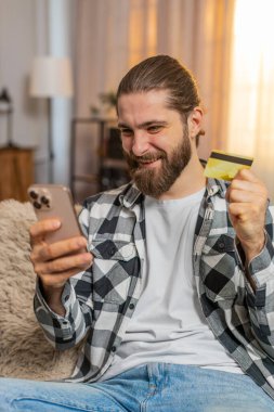 Genç adam evde telefon ve kredi kartıyla oturuyor, çevrimiçi alışverişi sonuçlandırırken toplamı doğruluyor. Kanepede oturan beyaz adam tamam, başarılı ödeme işlemlerine güvenle kafa sallıyor.