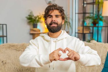 Ben de seni seviyorum. Oturma odasındaki kanepede oturan mutlu Hintli genç adamın portresi sevgiyi simgeler, kameraya kalp işaretini gösterir, romantik hisleri samimi ve olumlu duygular ifade eder. Minnettarlık.
