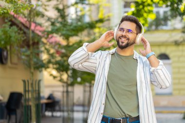 Kablosuz kulaklık takan mutlu beyaz adam dışarıda akıllı telefondan en sevilen enerjik müziği dinliyor. Şehir caddesinde yürüyen gülümseyen bir turist. Kasaba yaşam tarzları.