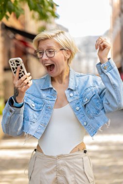 Aman Tanrım. Vay canına. Heyecanlı mutlu, mutlu, gözlüklü Avrupalı kadın dışarıda iyi haber kazanmak için akıllı telefon daktilo kullanıyor. Şehir caddesinde duran kız. Kasaba yaşam tarzları.