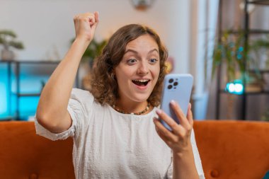 Mutlu genç bir kadın akıllı telefon daktilo kullanarak evet diyor, piyango galibiyetini kutluyor büyük ikramiye hedefi başarısını kutluyor iyi haber zaferi. Evdeki kız kanepede oturuyor. Yaşam Tarzları