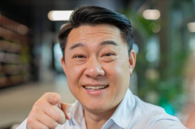 Mutlu Çinli işadamının yakın plan portresi el sıkışıyor selam veriyor, kameraya işaret ediyor, internetteki Webinar, uzaktan konuşma. Girişimci ofis lobisinde..