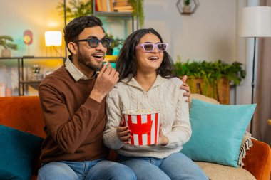 Üç boyutlu gözlüklü neşeli Hintli çift patlamış mısır yiyor ve evdeki oturma odasındaki kanepede oturmuş komedi filmi izliyor. Gülümseyen çeşitli aile hafta sonu eğlencesine gülüyor.
