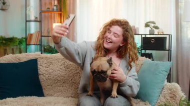 Beyaz saçlı genç bir kadın video görüşmesinde el sallıyor. Kanepede, gülümsüyor ve neşeli görünüyor. Kız pug dog 'u tutarken teması selamlıyor, yanında evcil hayvanla sevimli bir anı paylaşıyor..