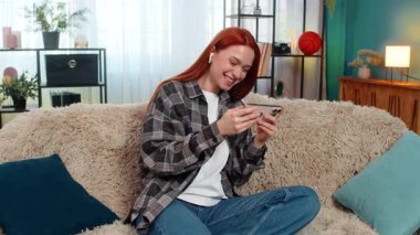 Genç bir kadın akıllı telefondan video izleyip kulaklık takıyor ve güçlü duygularla gülüyor. Kafkasyalı kızıl saçlı kız kanepenin üzerinde eğleniyor, neşe ve neşeyle tepki veriyor.