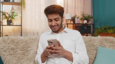 Kanepede oturan Hintli adam gülümsüyor, akıllı telefona bakarken başparmağını kaldırıyor, heyecan verici içeriğin tadını çıkarıyor. Arap adam olumlu tepki veriyor, onaylıyor, mutlu, kendinden emin ve tatmin olmuş hissediyor.