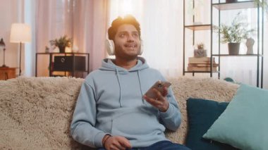 Kanepede kablosuz kulaklık takan akıllı telefondan müzik dinleyen genç Hintli adam favori pisti rahatlatmayı ve dans etmeyi seçiyor. İspanyol adam enerji ve neşe dolu ifadelerle ritmi seviyor..