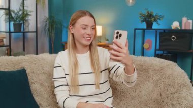 Beyaz bir kadın evdeki kanepede oturuyor, gülümsüyor, gülüyor, neşeli telefon görüşmesinde el sallıyor. Mutlu kız heyecan verici haberleri paylaşıyor, akıllı telefonu kullanıyor, evin içinde rahatça eğleniyor.