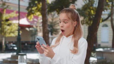 Akıllı telefon kutlaması yapan mutlu beyaz kadın iyi haber kazandı, piyango uygulaması zaferi, online açık hava. Şehir sokaklarında yumruklarını sıkan çekici bayan turist..