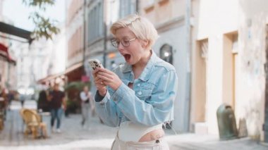 Kafkasyalı genç bir kadın cep telefonuyla kutlama yapıyor iyi haber kazanıyor, piyango büyük ikramiyesi kazanıyor, açık havada dağıtıyor. Şehir caddesinde zıplayan mutlu sarışın bayan turist. Kasaba yaşam tarzları.