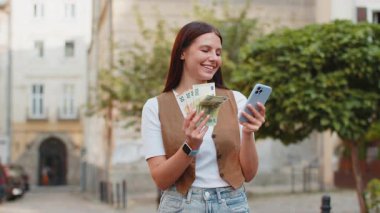 Mutlu genç bir kadın, euro nakit para sayıyor, akıllı telefon hesap makinesi uygulaması kullanıyor, planlanmış tatil hediyeleri için gelirden memnun. Şehir merkezinde güneşli bir sokakta duran güzel bir kız. Şehir yaşam tarzları açık havada