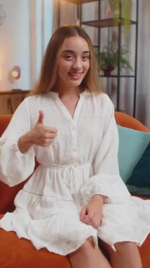 Gülümseyen beyaz kadın portresi onaylayıcı bir şekilde kameraya bakıyor işaret gibi, olumlu geri bildirim, iyi haber. Evdeki oturma odasındaki kanepede oturan mutlu kız. Dikey