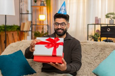 Evdeki Hintli adam mutlu bir şekilde doğum günü hediyesini açarken koltukta gülümseyerek ve heyecanla oturuyor. Arap adam şaşırmış ve neşeli görünüyor. Beklenmedik bir hediye ve özel bir kutlama anı karşısında heyecanlanıyor.