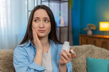 Şiddetli diş ağrısından muzdarip genç hasta kadın, yanağını tutuyor, yorulmadan ağrı kesici haplar arıyor. Kafkasyalı hasta kız diş ağrısından bitkin düşmüş.