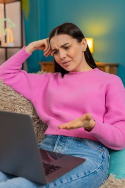 Evdeki genç kadın dizüstü bilgisayar ekranına bakmaktan memnun değil. Üzücü bir ifadeyle baş aşağı duruyor. Kanepede oturan beyaz kız yorgun, iş yükü ve olumsuz duygular yüzünden hayal kırıklığına uğramış.