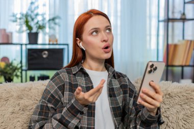 Evdeki genç kadın mobil uygulama kullanarak yeni bir dil öğreniyor ve kelimeleri odaklanma ve neşeyle yüksek sesle tekrarlıyor. Kanepedeki beyaz saçlı kız eğitim sürecinin tadını çıkarıyor. Telaffuz ve konuşma pratiği yapıyor.