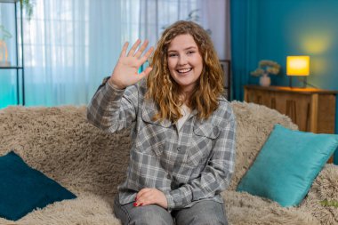 Evdeki genç kadın sıcak bir şekilde gülümsüyor ve sıcak ve neşeli bir şekilde merhaba demek için el sallıyor. Kanepede oturan beyaz saçlı kız neşe, hoş geldin jesti ve olumlu duygusal bağlarını dile getiriyor.