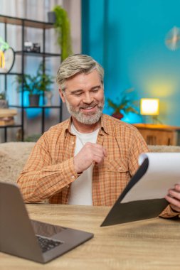 Olgun bir adam evde oturmuş önemli belgeleri inceliyor ve dizüstü bilgisayar kullanıyor. Orta yaşlı bir adam başarılı bir işten aldığı memnuniyetle gülümser, verimlilik ve başarıyı gösterir. Dikey