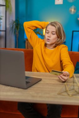 Beyaz çocuk evde dizüstü bilgisayarından online alışveriş yapıyor. Kredi kartı bilgilerini girmek, yeni oyun ögesine tıklamak, koltuk masa otururken, satın almaktan memnun olmak