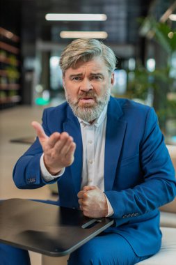 Çatışma Anlaşması 'nda. Olgun kızgın patron iş adamı webcam çevrimiçi video görüşmesi yapıyor, kameraya bakarak iş arkadaşlarıyla konuşuyor. Takım elbiseli beyaz adam modern ofis lobisinde oturuyor..