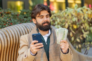 Akıllı telefon kullanan ve paralı banknotları sayan mutlu Hintli işadamı şehir merkezindeki bankta oturuyor. Başarılı iş kariyeri, piyango kazananı, büyük gelir, zenginlik