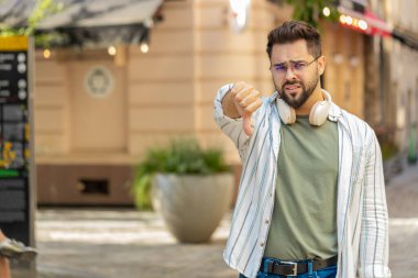 Hoşlanmamak. Kafkasyalı genç adam işaret dilini gösteriyor, hoşnutsuzluğunu dile getiriyor, tatminsiz kötü işler yapıyor, açık havada hata yapıyor. Şehir caddesinde duran hoşnutsuz turist..