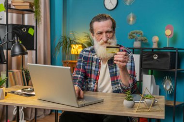 Merkez ofisteki online alışveriş ödemeleri için banka kartı ve dizüstü bilgisayar kullanan Kafkasyalı kıdemli bir adam. İş anlaşması. Gülümseyen büyükbaba, E-bankacılık uygulaması ile serbest alım yapıyor..